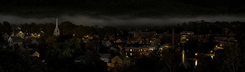 An illustration of a small mill town at night in a river valley, with low fog, lit windows and a white steeple